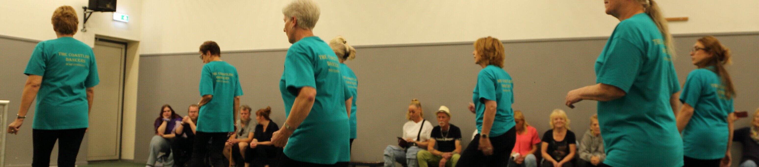 Seniorengroep danst in blauwe shirts tijdens een line-dance demonstratie in een sportzaal, met publiek op de achtergrond.