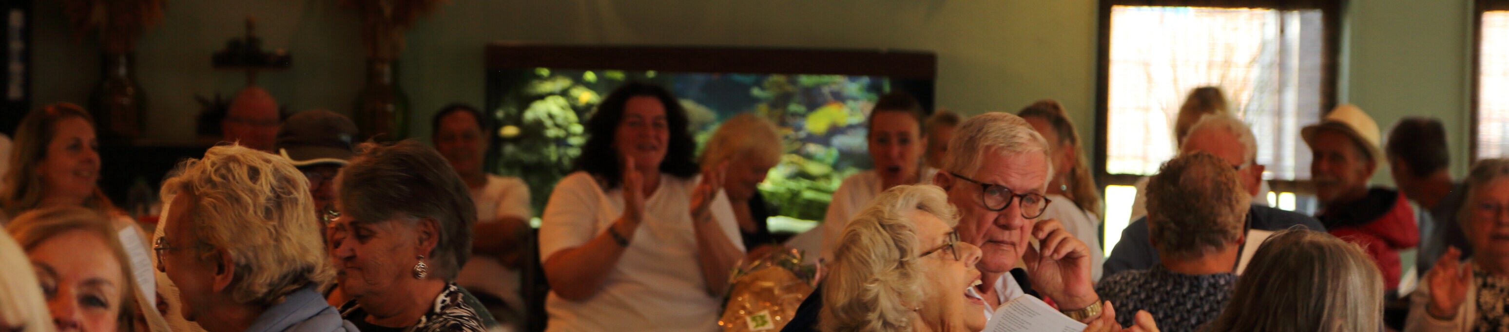 Senioren zingen en lachen samen tijdens een feestelijke bijeenkomst bij Welzijn Scheveningen, onder slingers met het getal 50.